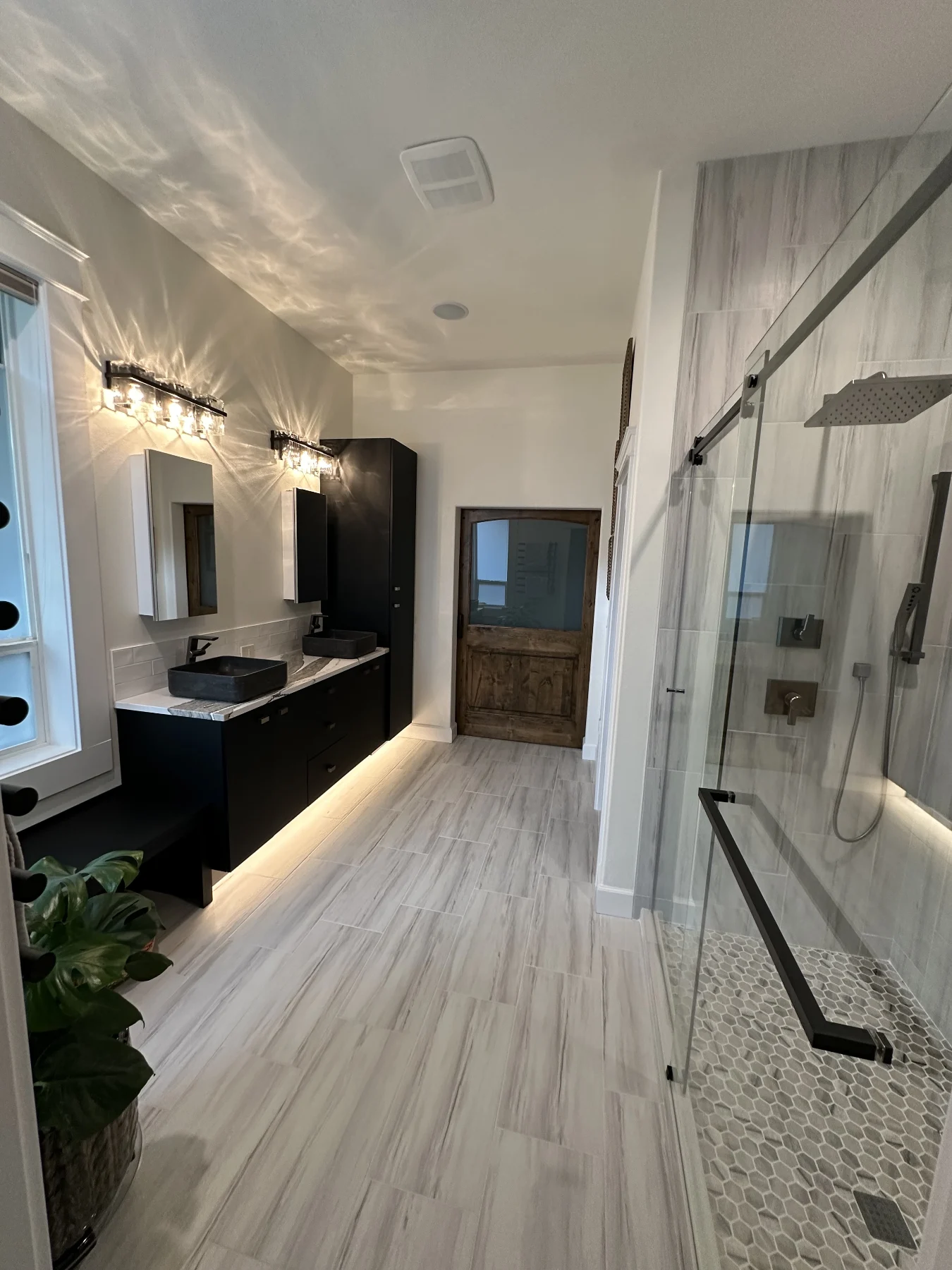 Bathroom view along a black double vanity toward a wood door with a glass shower on the right