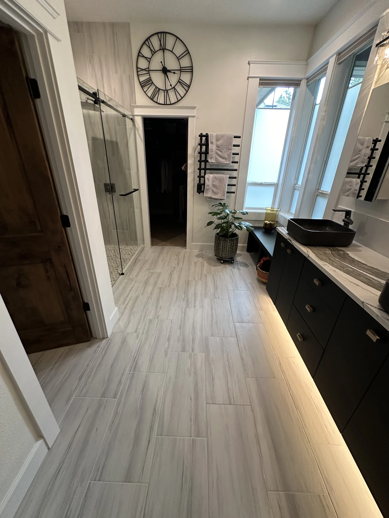 Bathroom with pale floor tile, a black vanity, tall windows and a glass shower