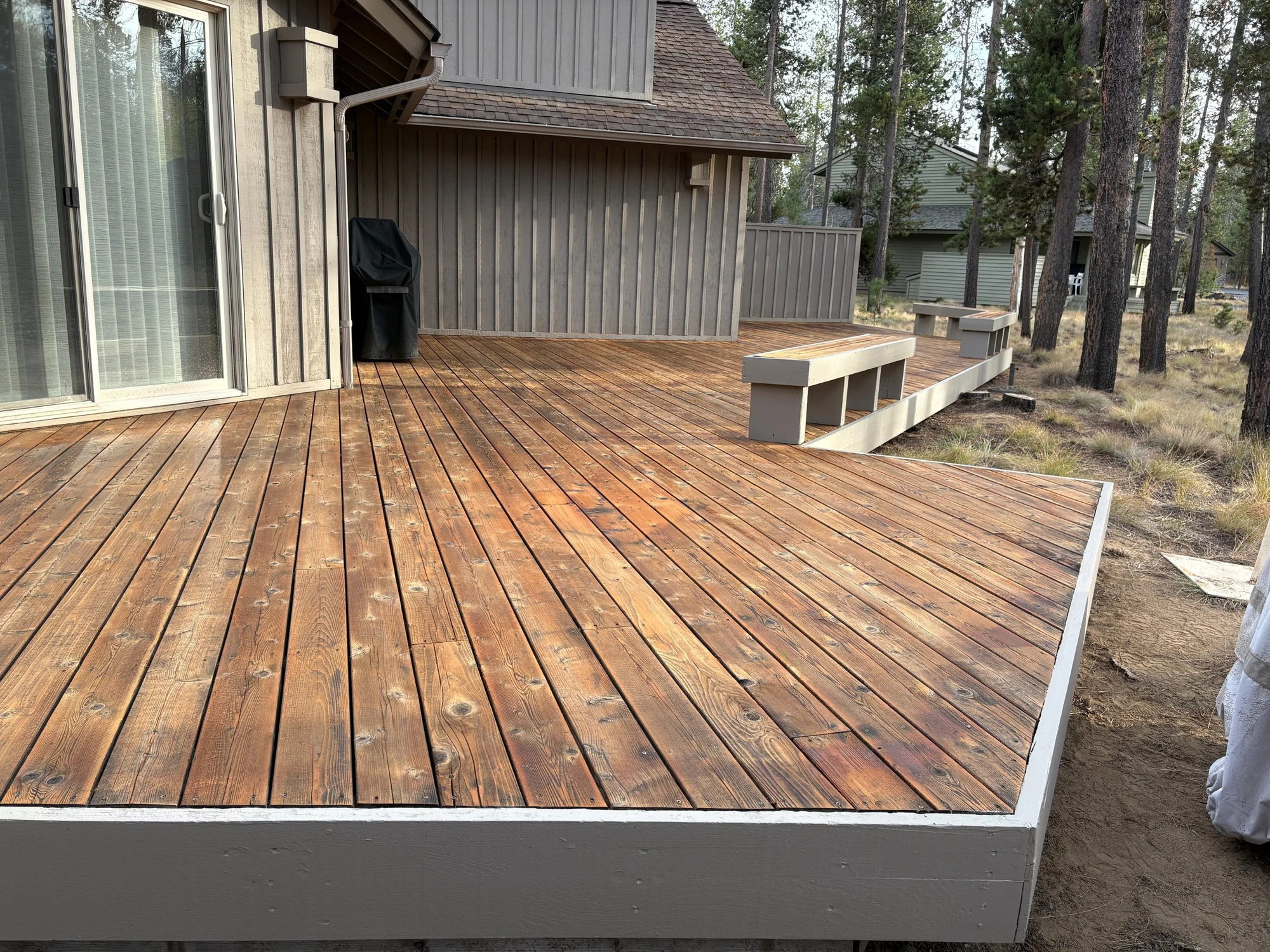 Cedar deck with a built-in bench beside a gray home among pine trees