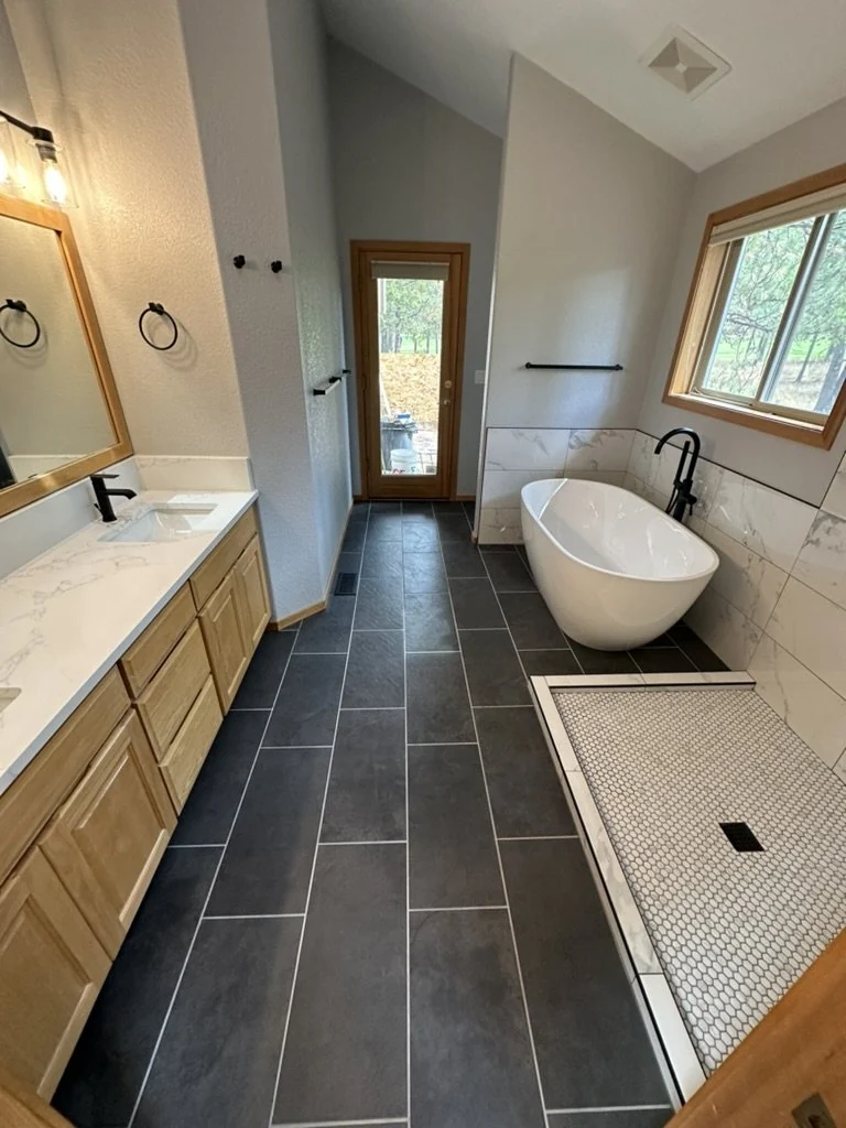 White freestanding tub, pale wood vanity and dark floor tile in a remodeled bathroom